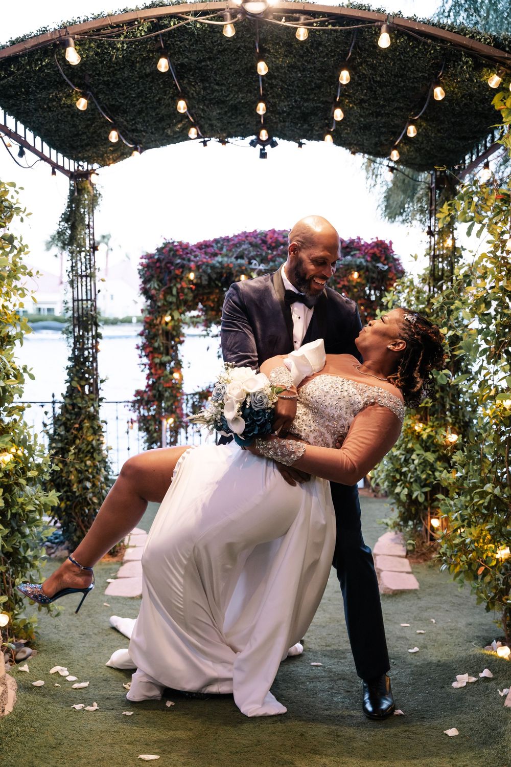 A romantic couple shares an intimate dance dip moment under a garden arbor decorated with string lights and flowers.