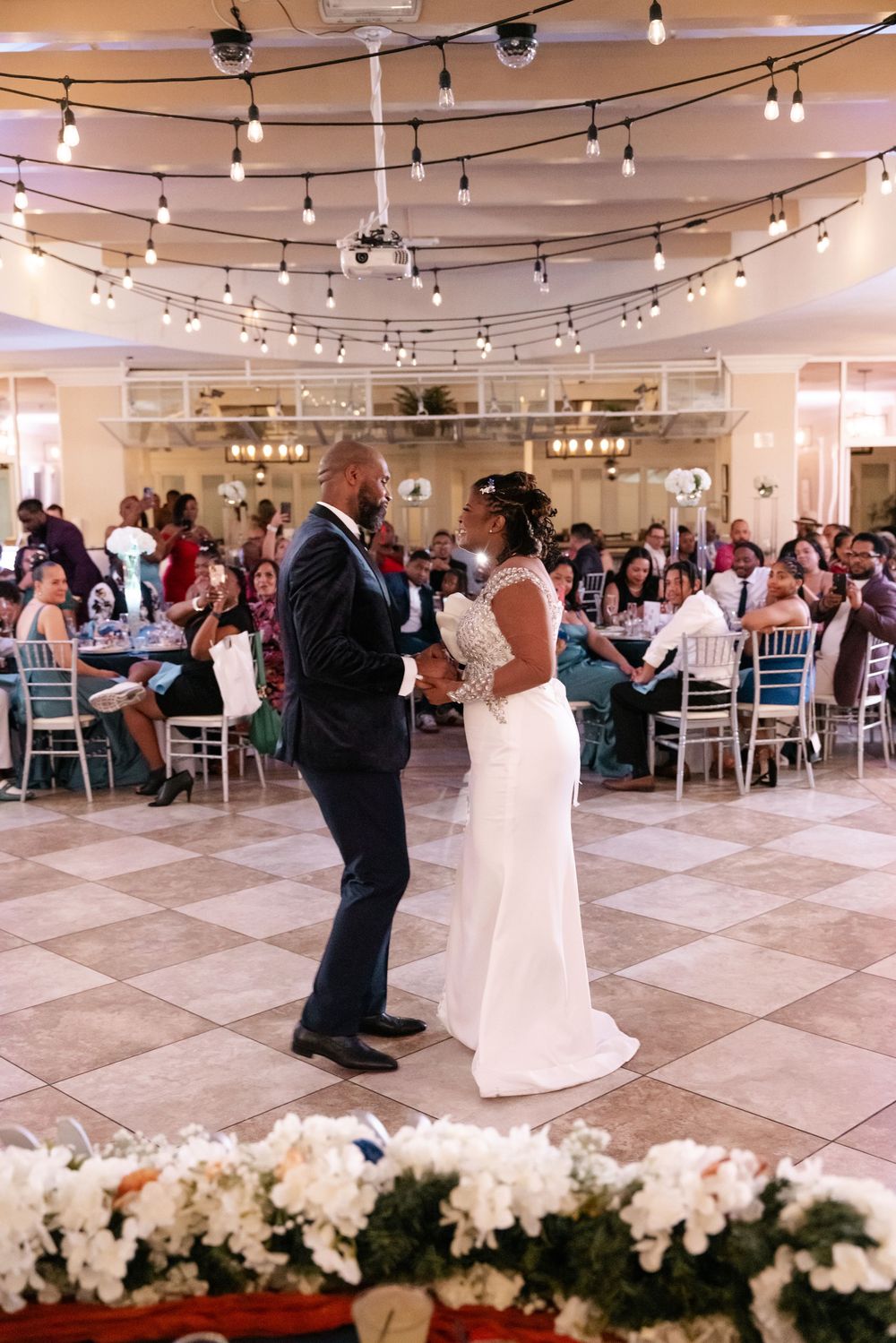 A couple shares their first dance in an elegant ballroom with string lights overhead and guests seated at round tables.