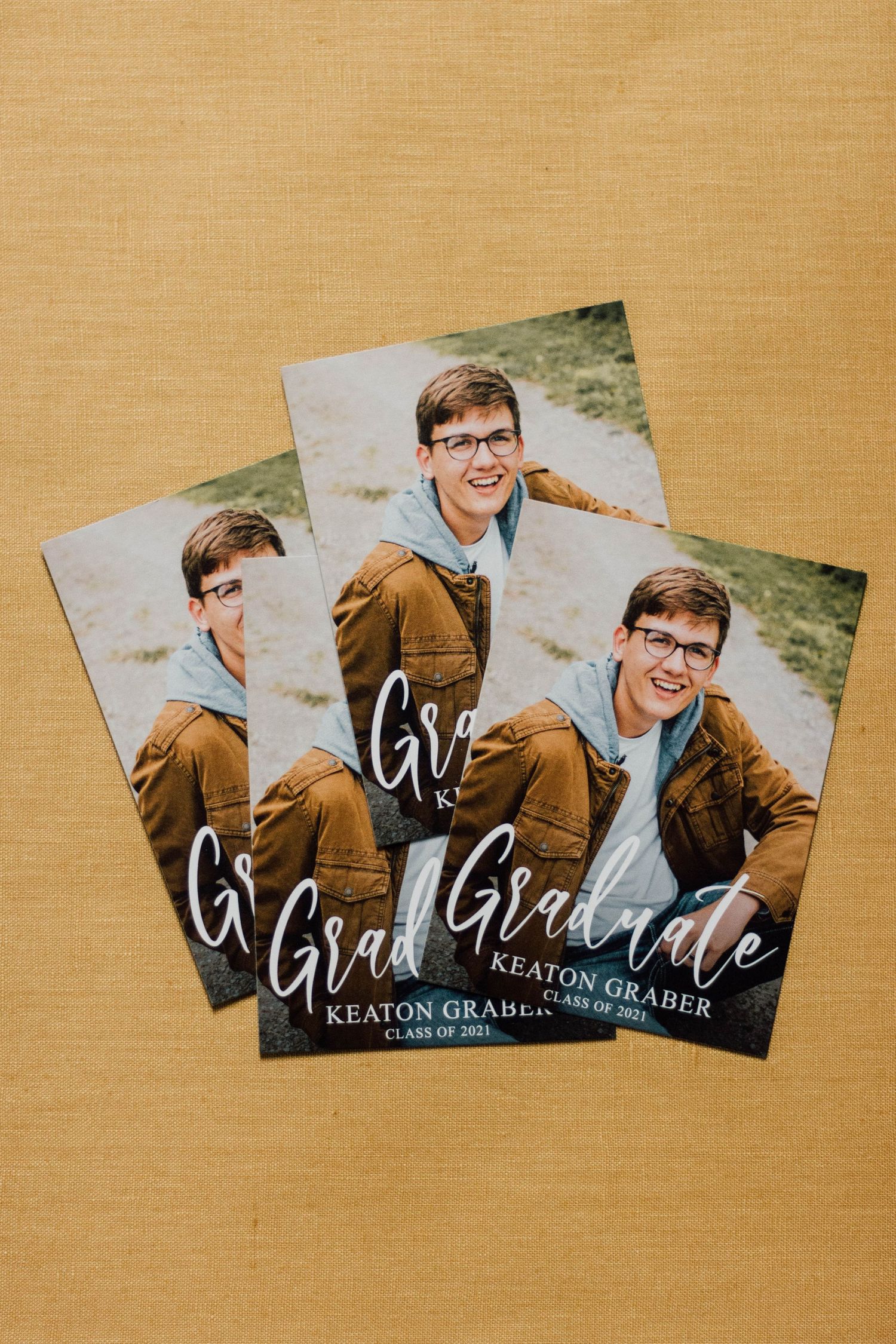 Graduation announcement cards featuring brown and white text arranged in a fan layout on a tan background.