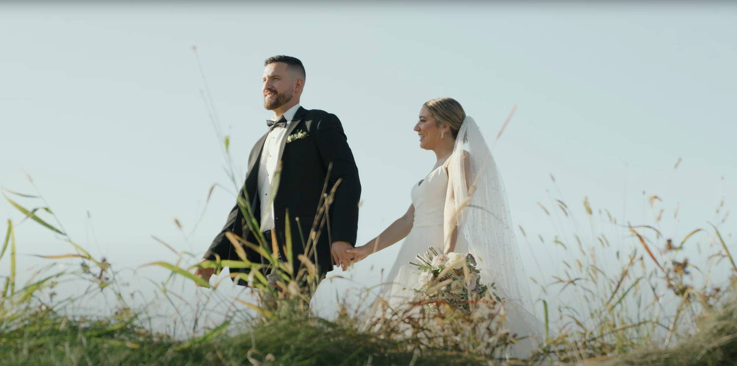 A couple in wedding attire walks through tall grass in a meadow at sunset.