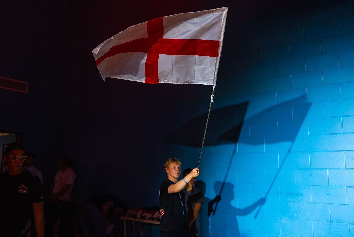 Person waving an England St George's Cross flag in a dimly lit blue-walled venue during a sporting event.