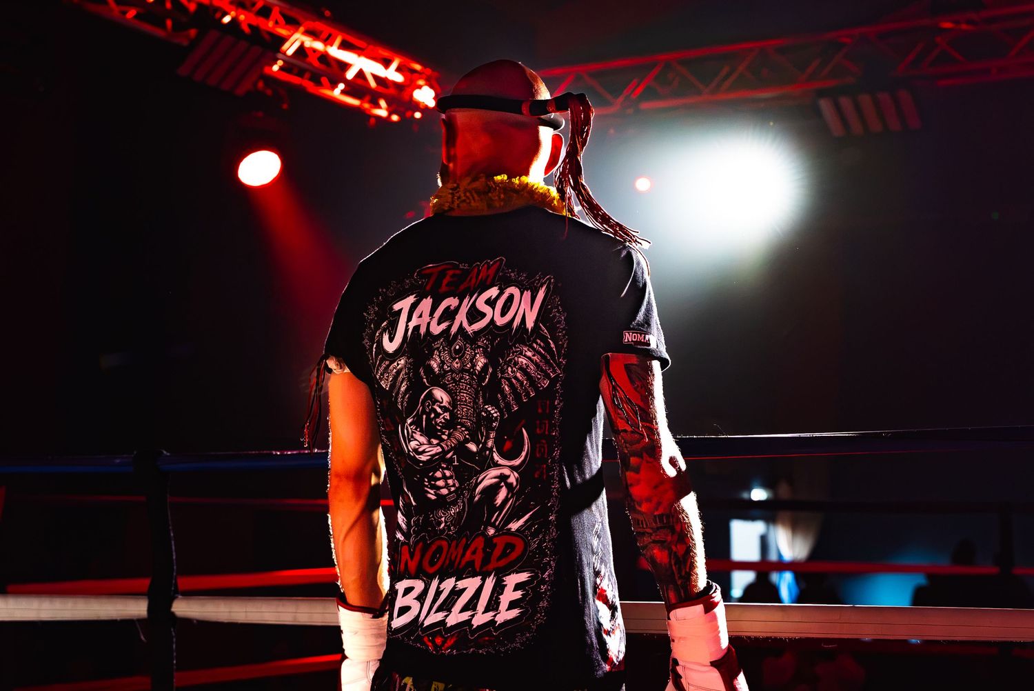 Wrestler in Jackson Bizzle graphic tee stands ringside under dramatic red stage lighting.
