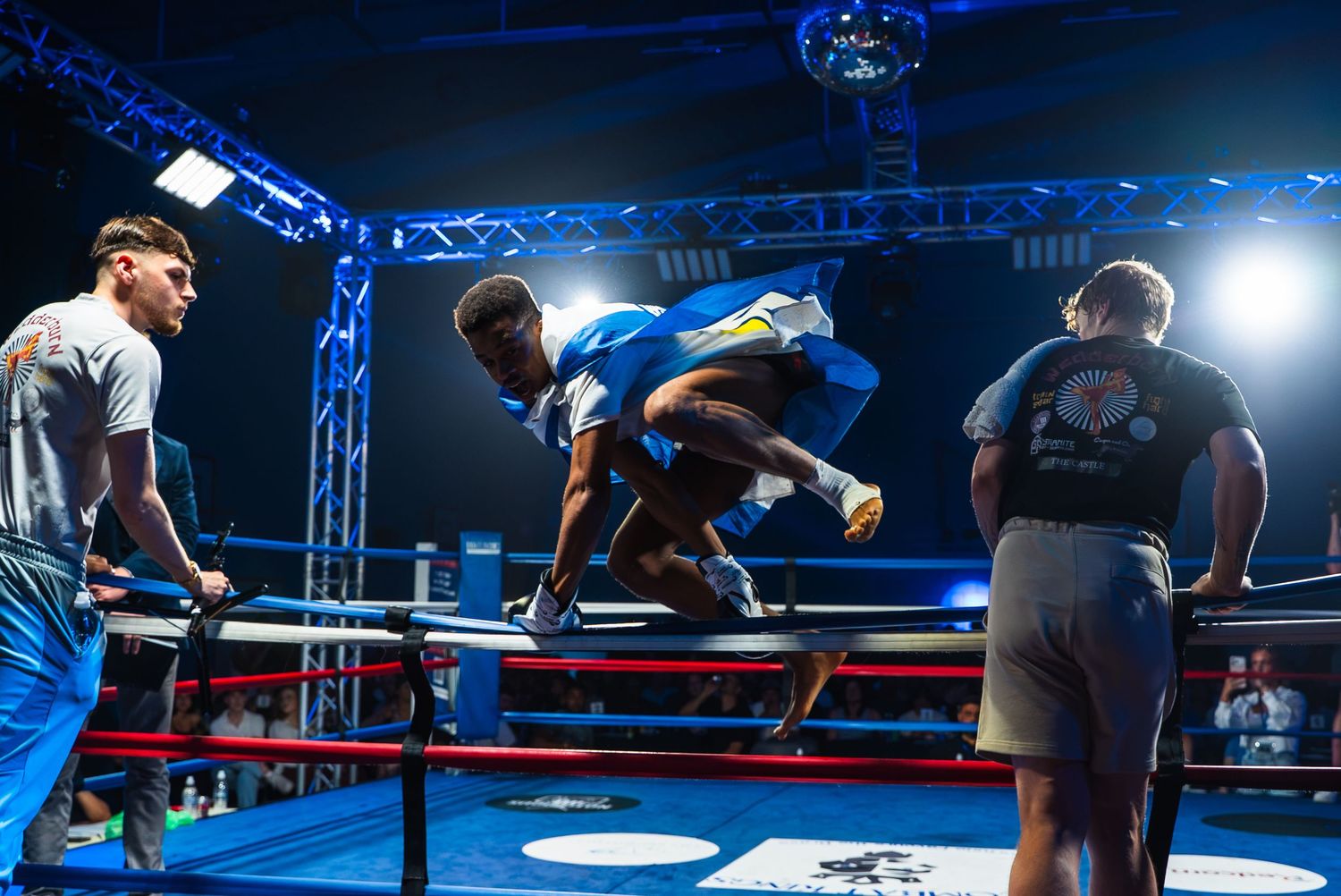 Boxer leaping into a boxing ring during a professional fight event under bright studio lights.
