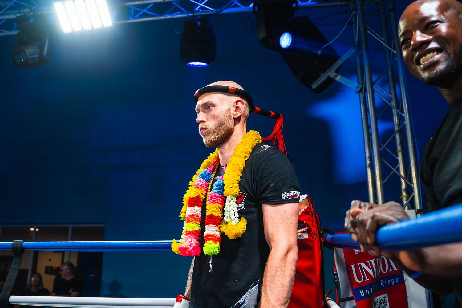 A boxer wearing a colorful floral lei stands confidently in a boxing ring under bright lights before a match.