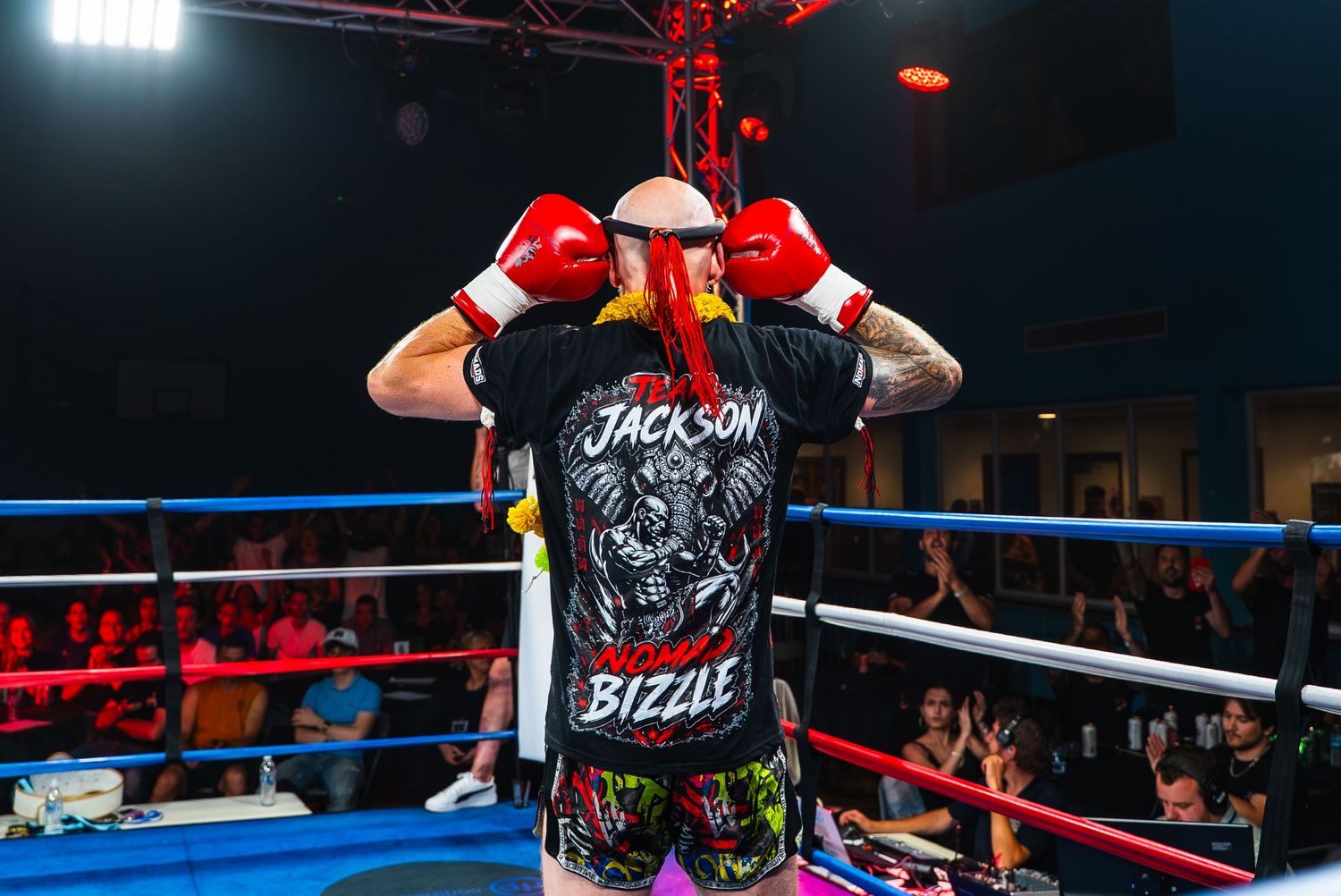 Boxer named Jackson Bizzle stands in a fighting ring wearing red gloves and black graphic tee, facing away from camera.