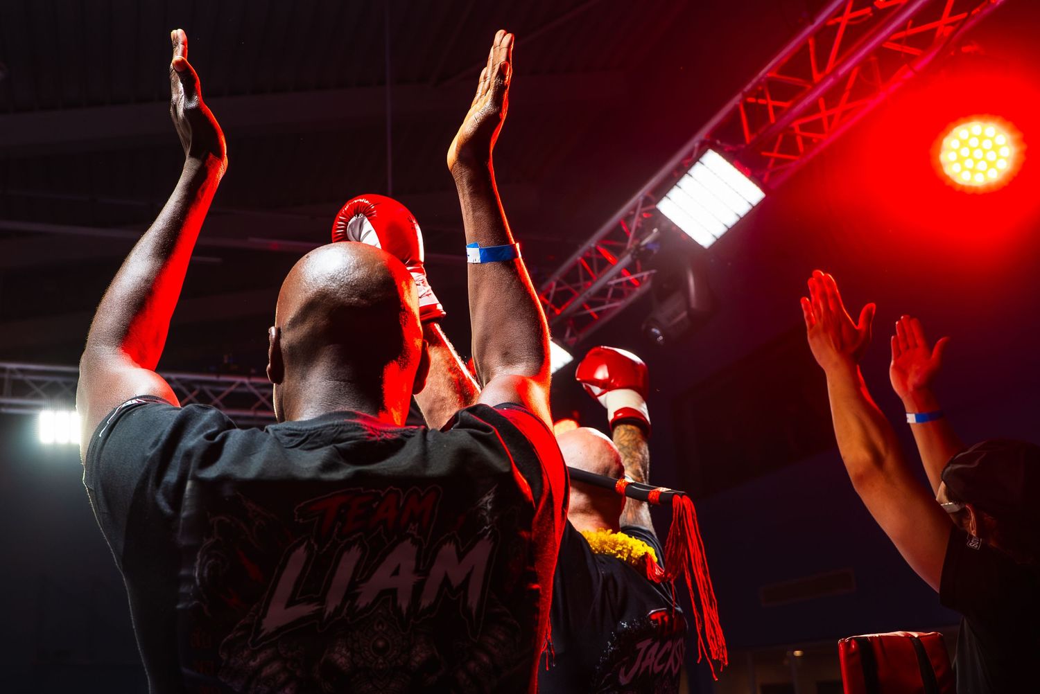 Fighters raise hands under red stage lights at a boxing event, one wearing a black shirt reading LIAM.