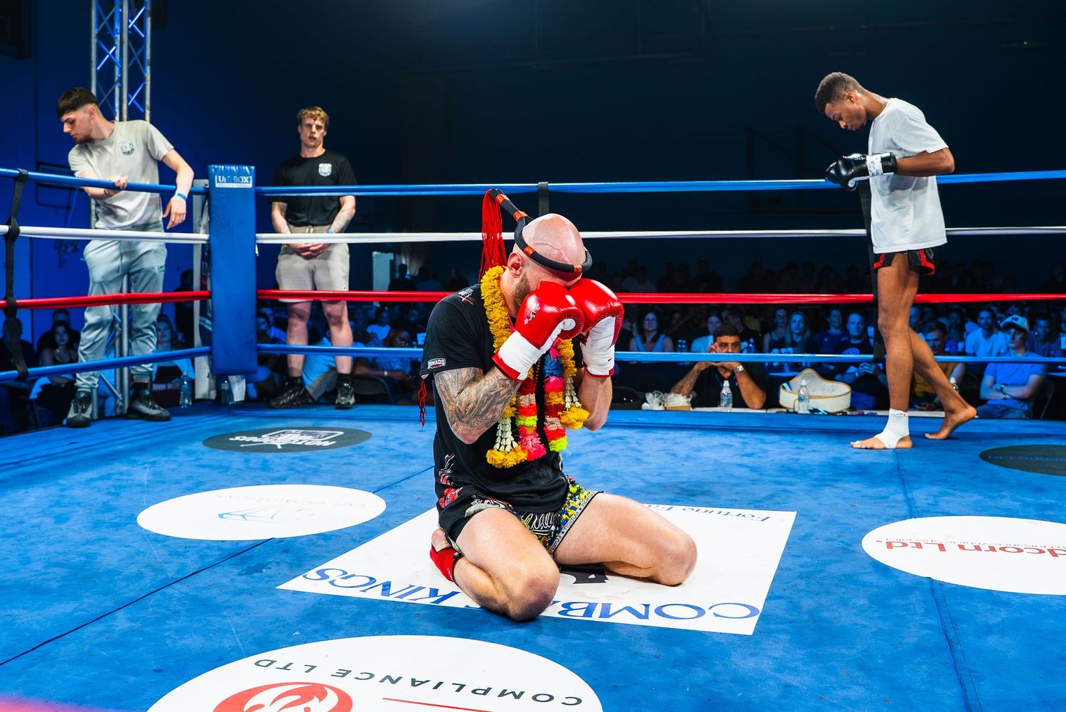 Boxer kneels on blue ring canvas celebrating victory, holding red scarf, during professional boxing match indoors.