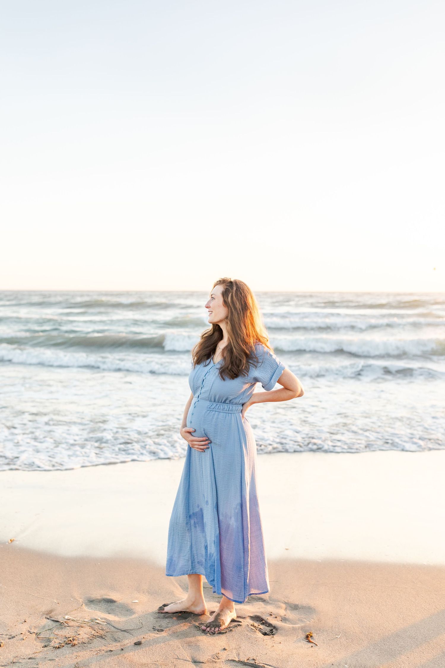 Someone in a flowy light blue dress twirls joyfully on a sandy beach at sunset with ocean waves in the background.