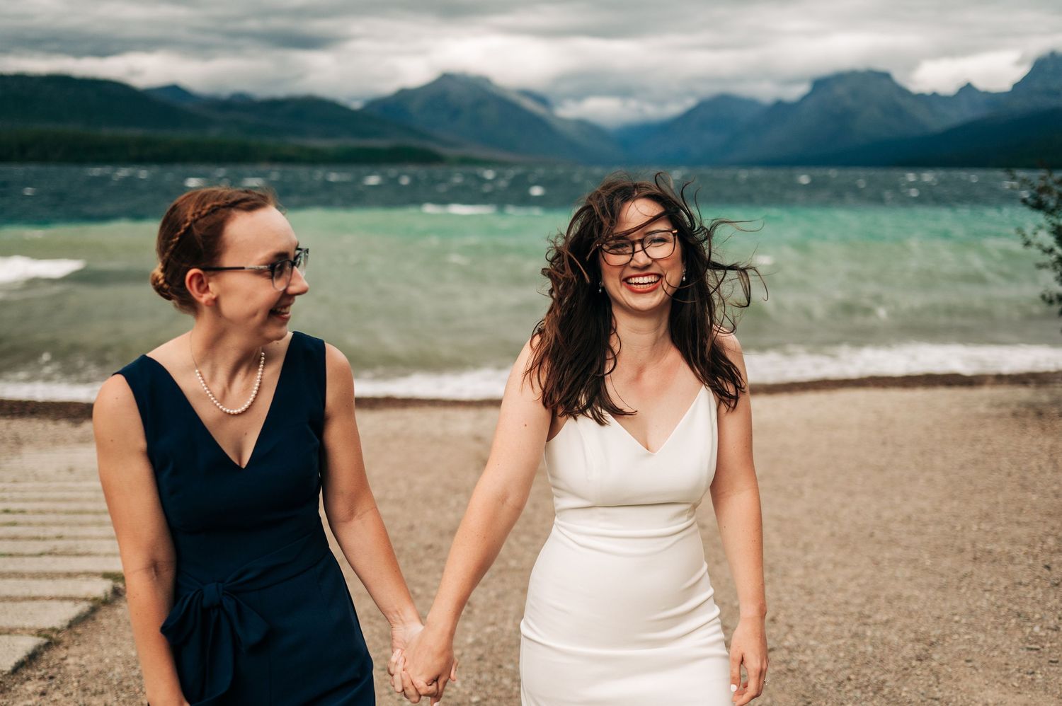 A couple walking hand in hand along a sandy beach with mountains and turquoise water in the background on a cloudy day.