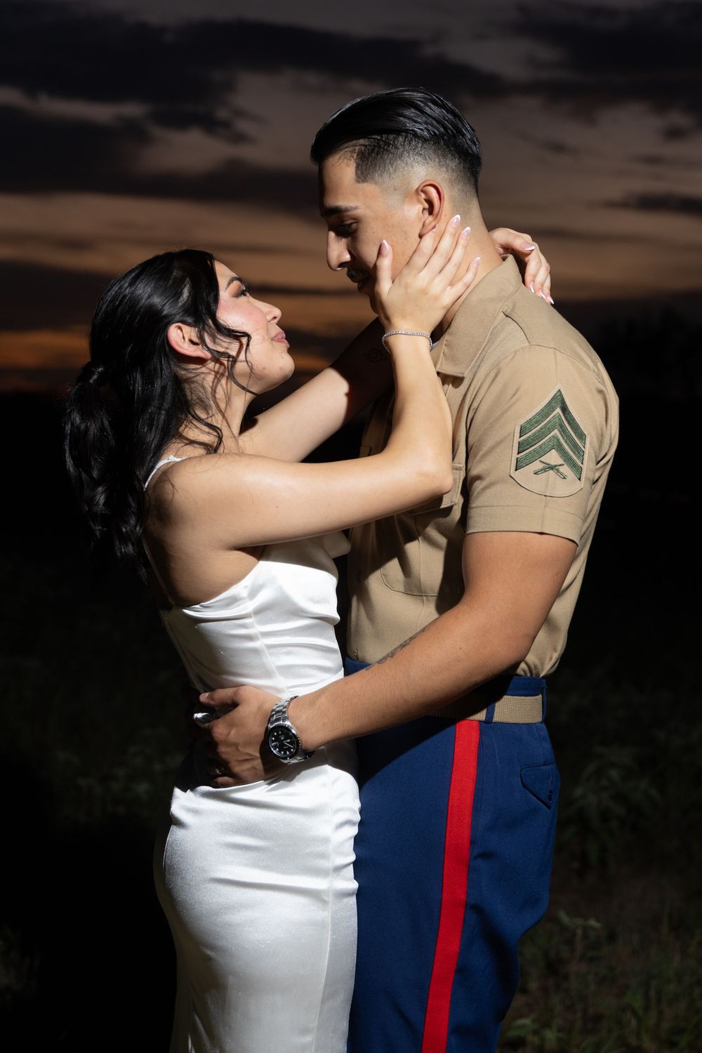 A couple in formal attire shares a romantic embrace at sunset in an outdoor setting.