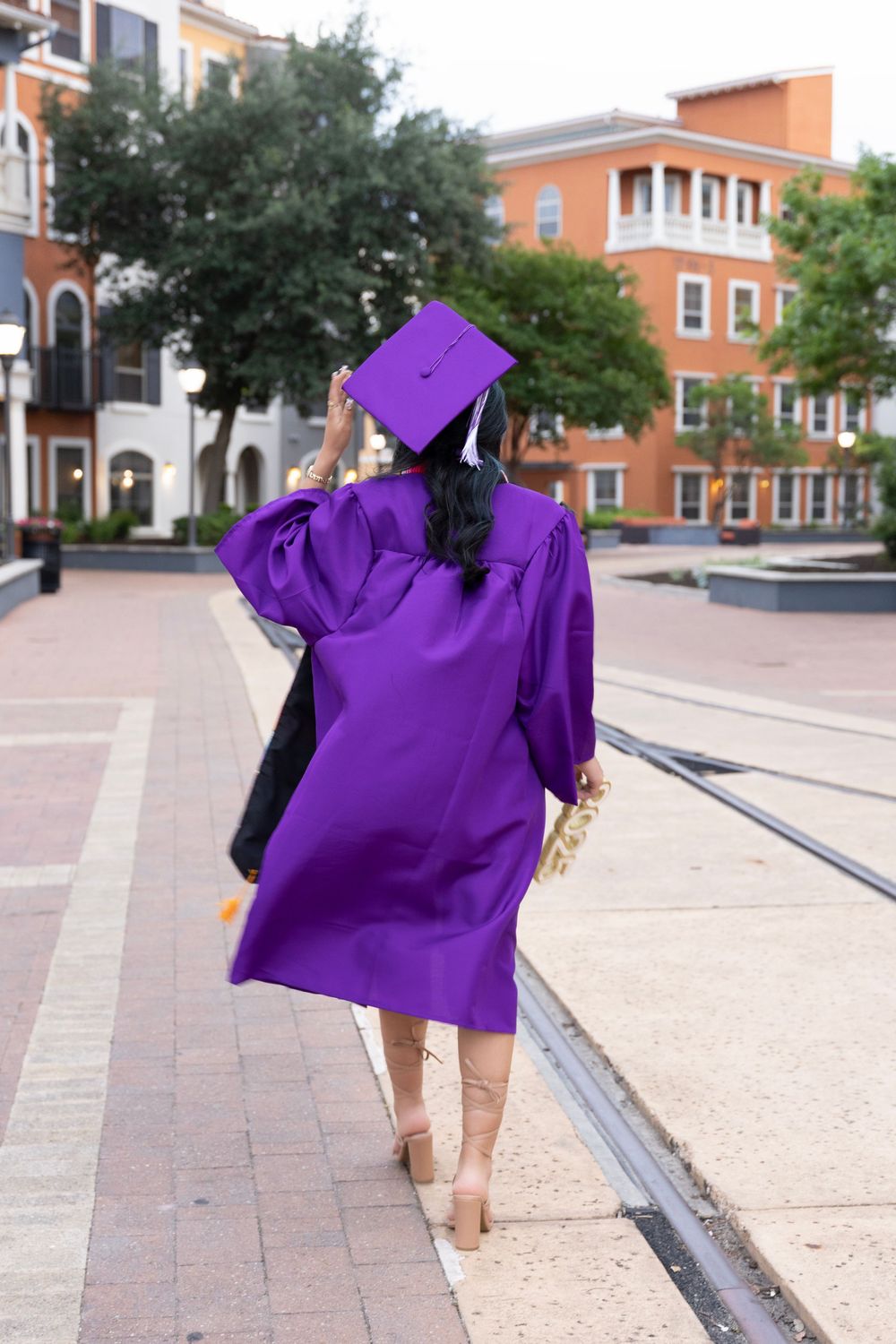 A person in a vibrant purple graduation gown walking on brick pavement near red brick buildings on a university campus.