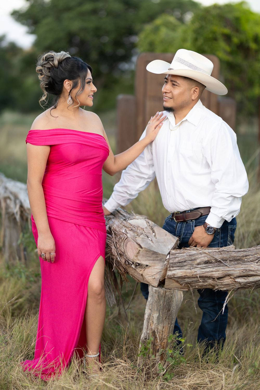 A couple in pink dress and western attire pose romantically against a rustic wooden log in a rural outdoor setting.