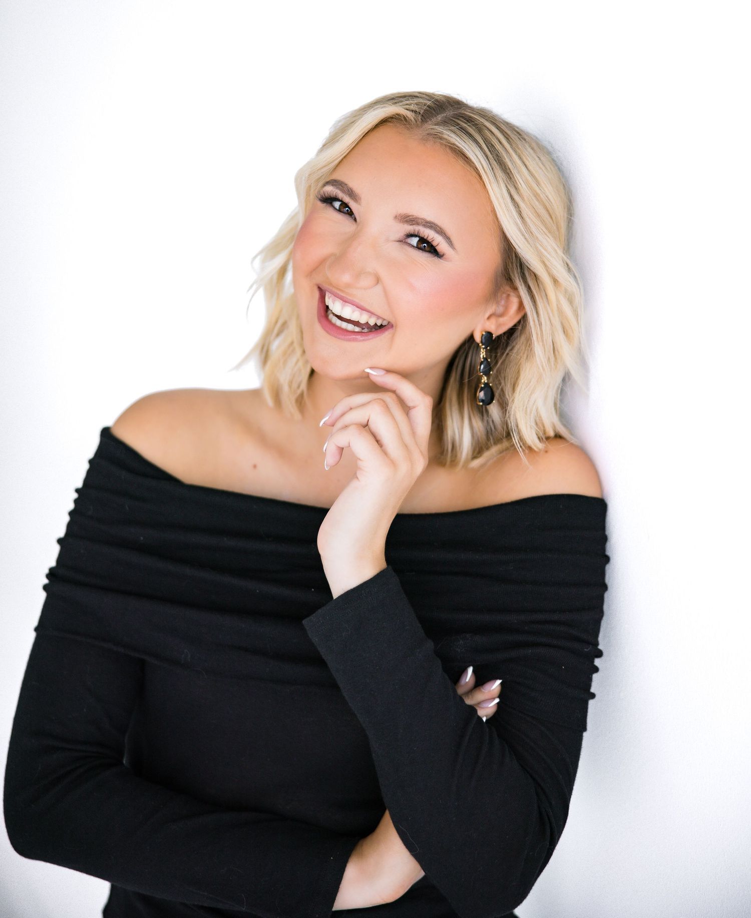 A woman in a black off-shoulder top smiles while posing against a white background in a beauty photoshoot series.