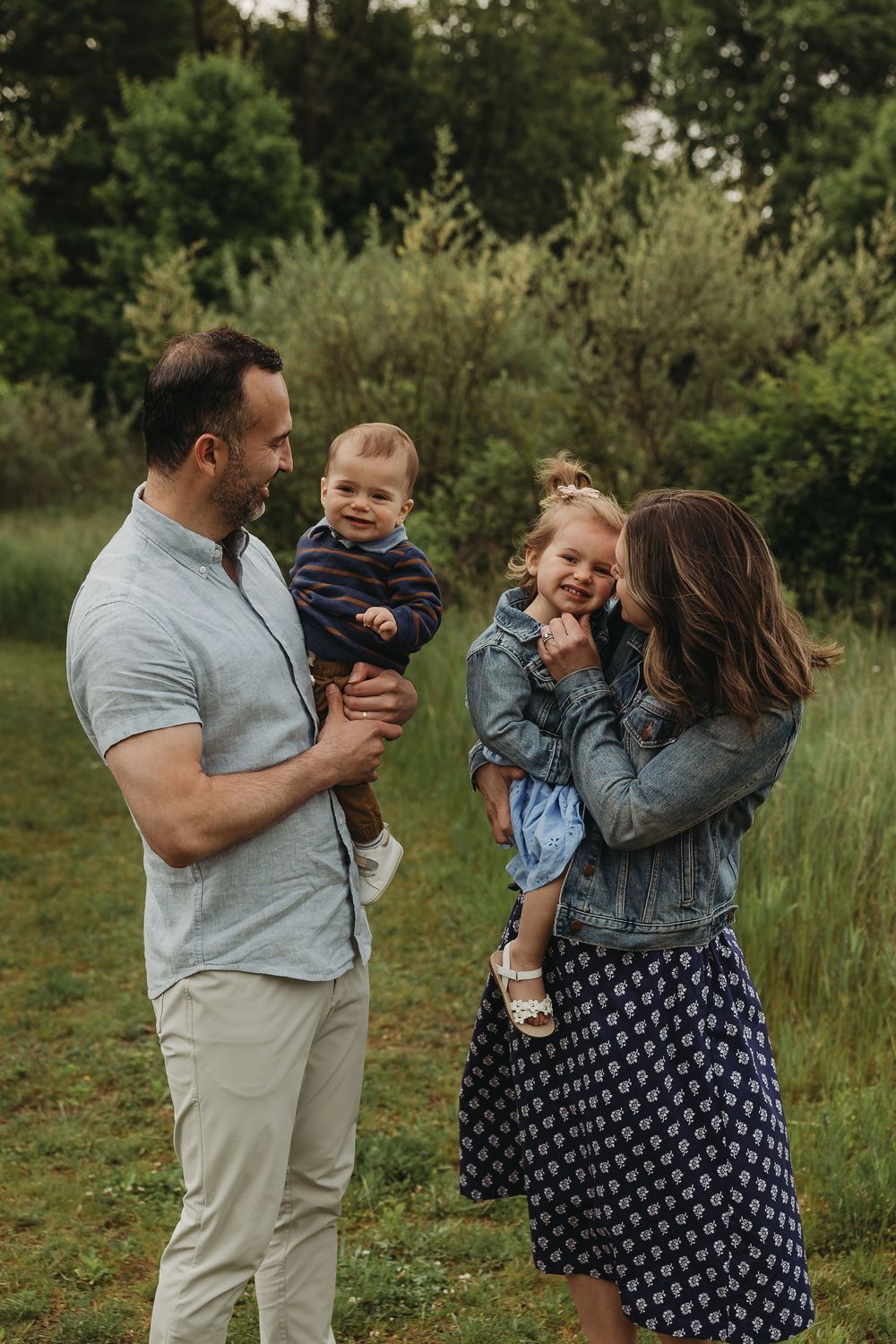 A loving family of four enjoys a candid outdoor moment together in a natural grassy setting.