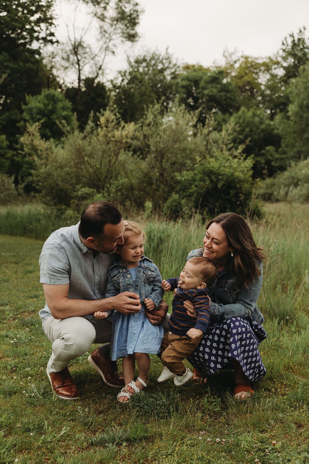 A candid outdoor family moment captured in a grassy field with trees in the background during golden hour.