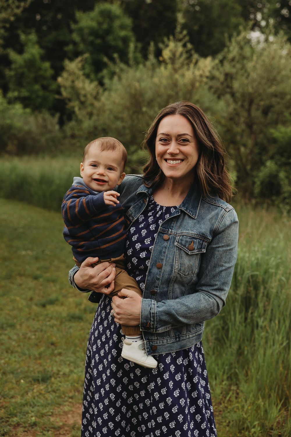 A parent holds a baby while wearing a denim jacket and patterned dress against a green outdoor background.