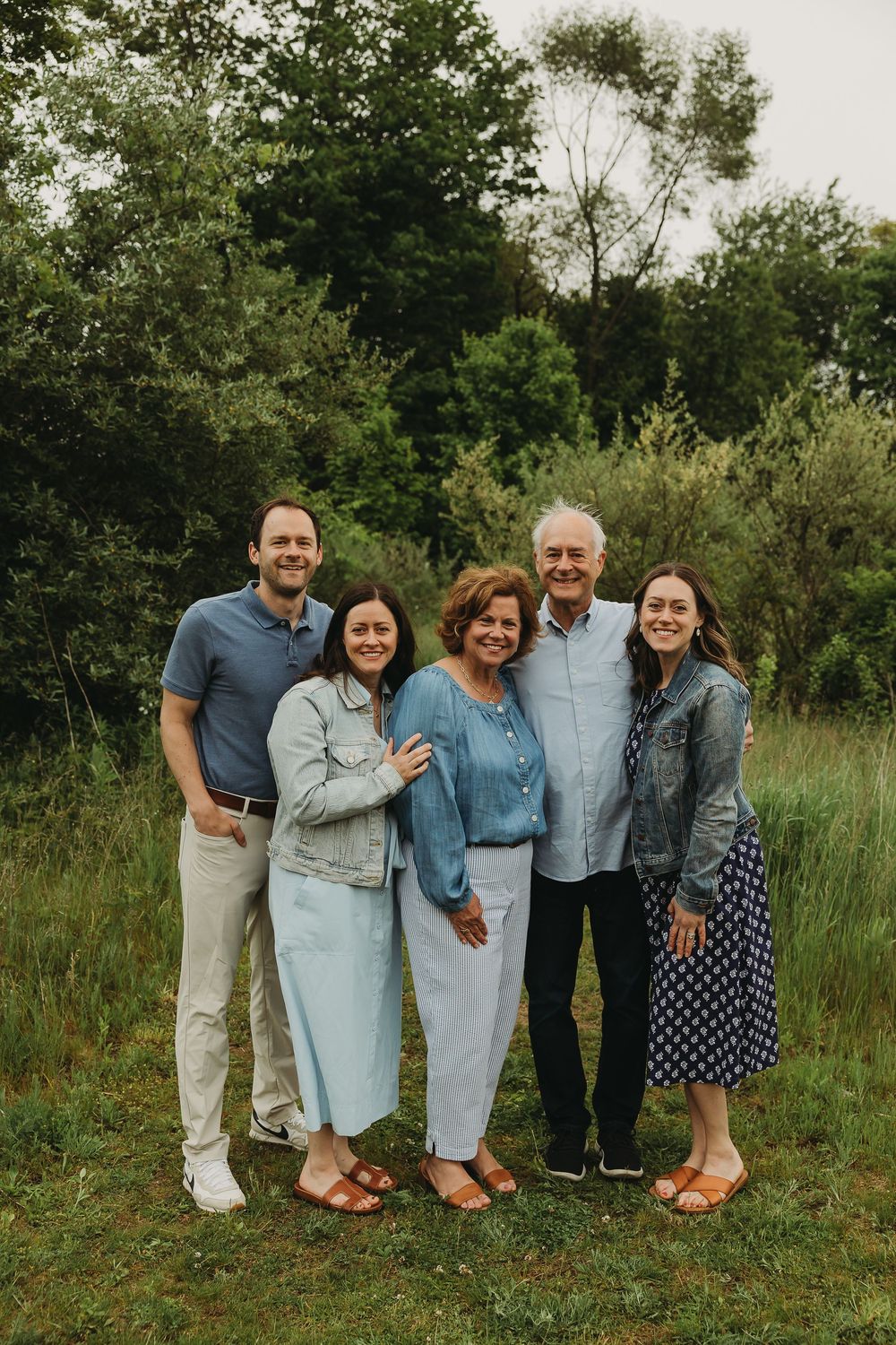 A group of five people stand together outdoors in front of green trees and foliage on a summer day.