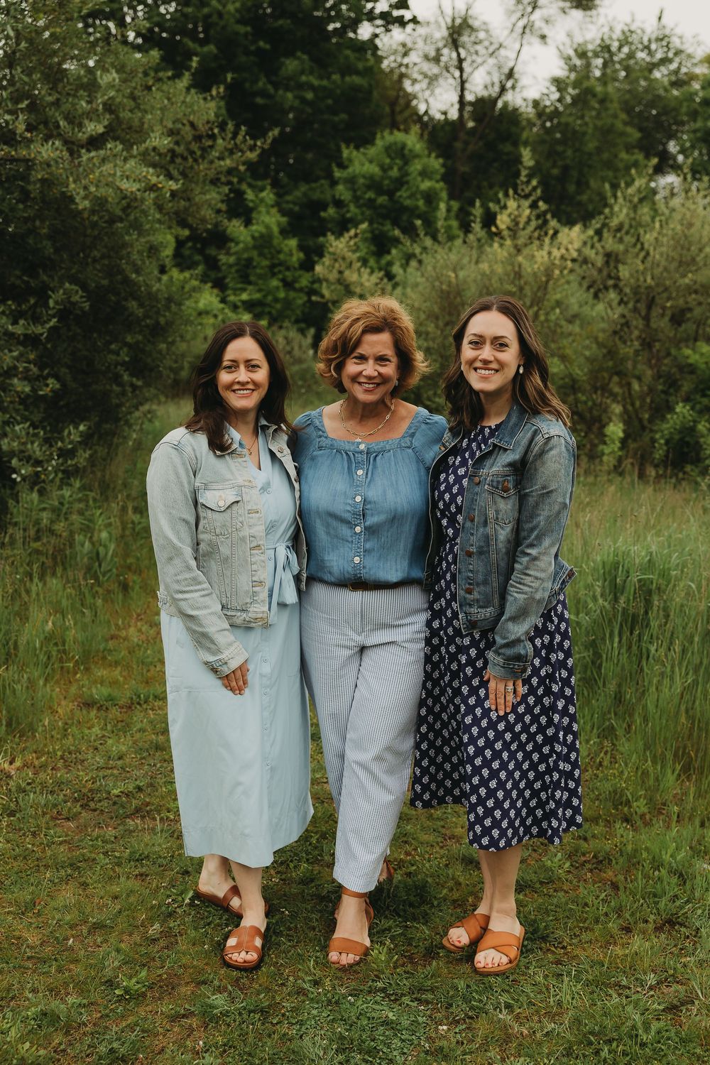 Three people wearing casual denim and blue outfits pose together on green grass in a natural outdoor setting.
