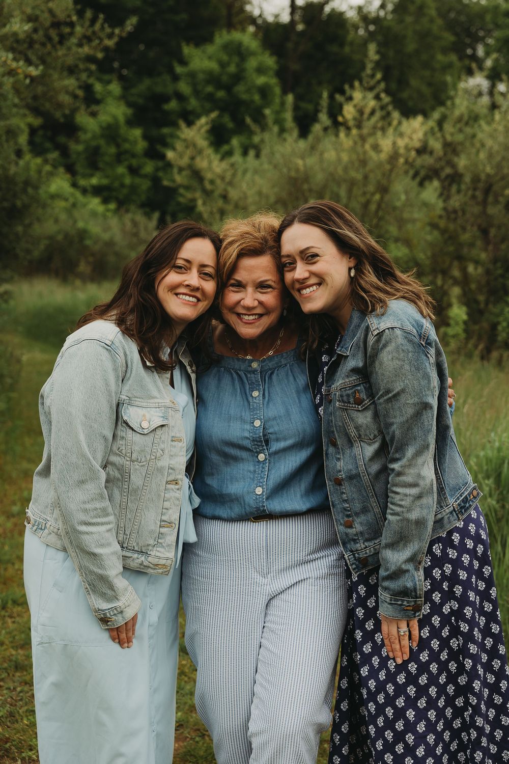 Three friends wearing denim jackets and casual outfits embrace and smile together in a grassy outdoor setting.