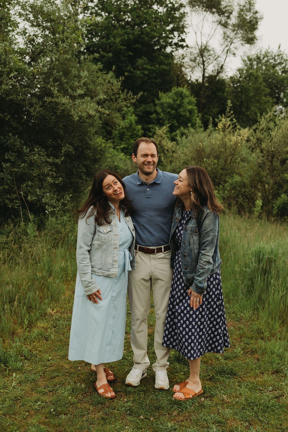 Three people stand together outdoors in casual attire smiling on grassy ground with green foliage in the background.
