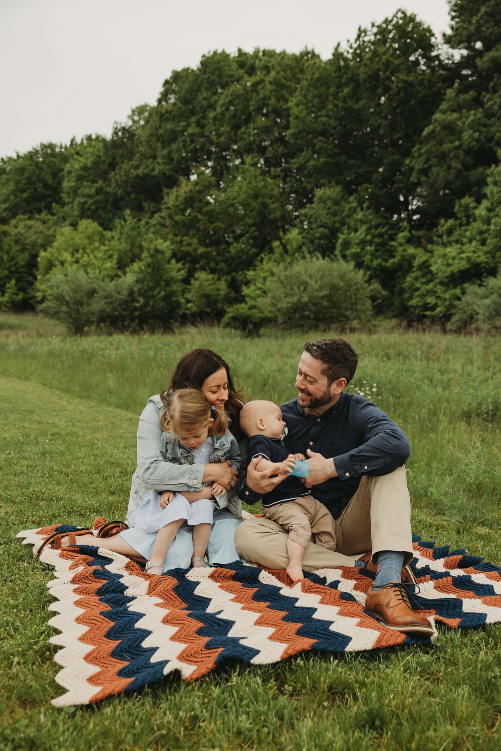 Family enjoying a picnic together on colorful zigzag patterned blanket in grassy field surrounded by green trees.