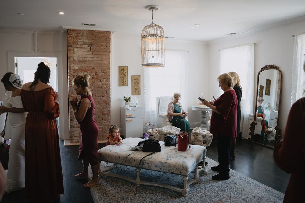 Bridesmaids in burgundy dresses gather in elegant bridal suite with chandelier and exposed brick wall.