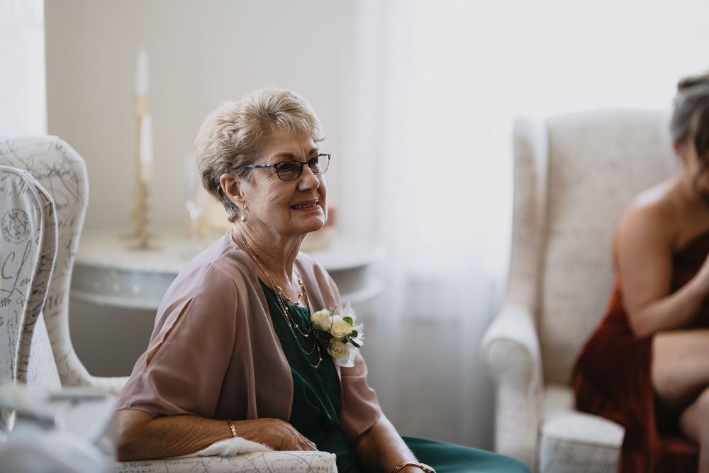 Elderly woman wearing glasses and a corsage smiles warmly at a wedding ceremony while seated in a chair.