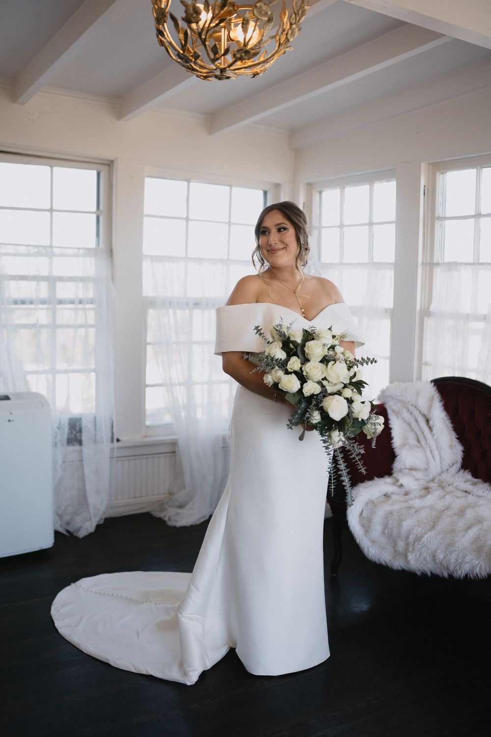 Bride in off-shoulder white gown holding white rose bouquet stands in sunlit room with elegant chandelier.