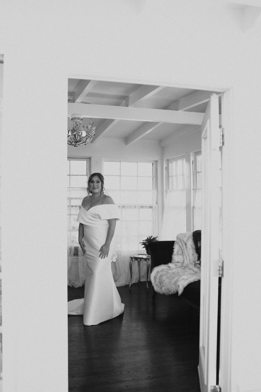 Bride in elegant off-shoulder mermaid wedding dress standing in doorway of bright room, black and white photo.