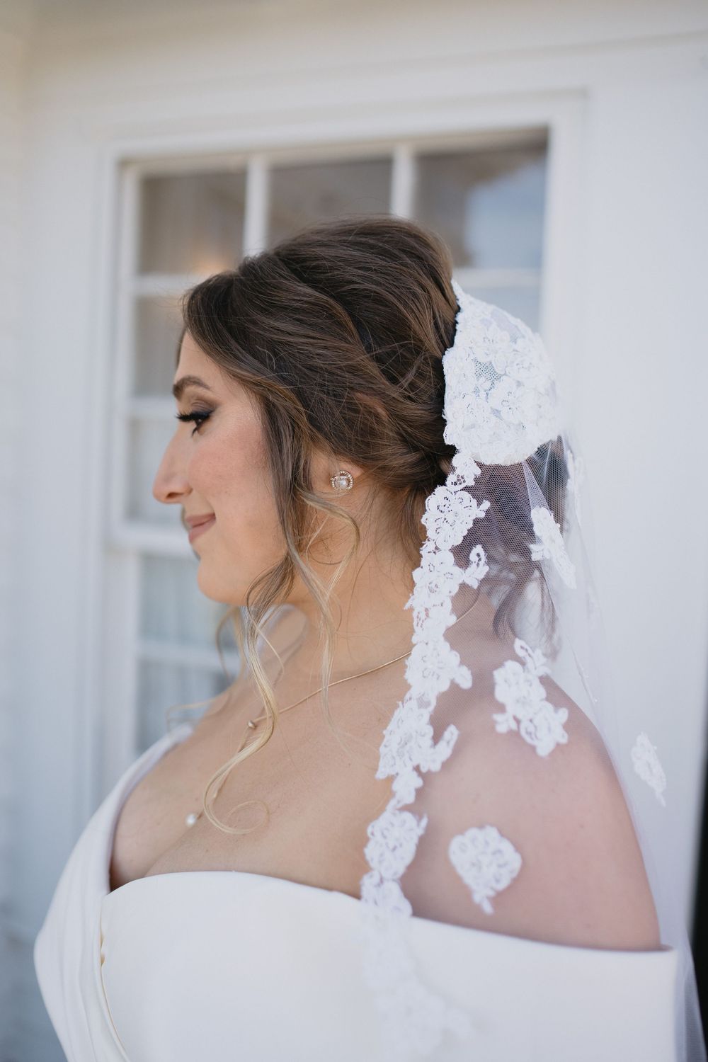 Bride in off-shoulder gown with lace veil and updo hairstyle, smiling in profile view outdoors.