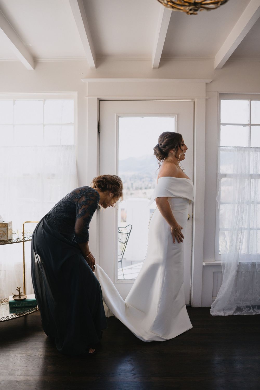 A bride in an off-shoulder white gown stands as a woman in navy adjusts her train in a bright, elegant room. at Manor House Colorado