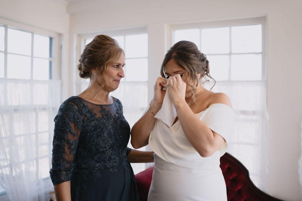 Emotional bride in white gown wiping tears as older woman in blue lace dress comforts her before wedding ceremony. By cinematic destination