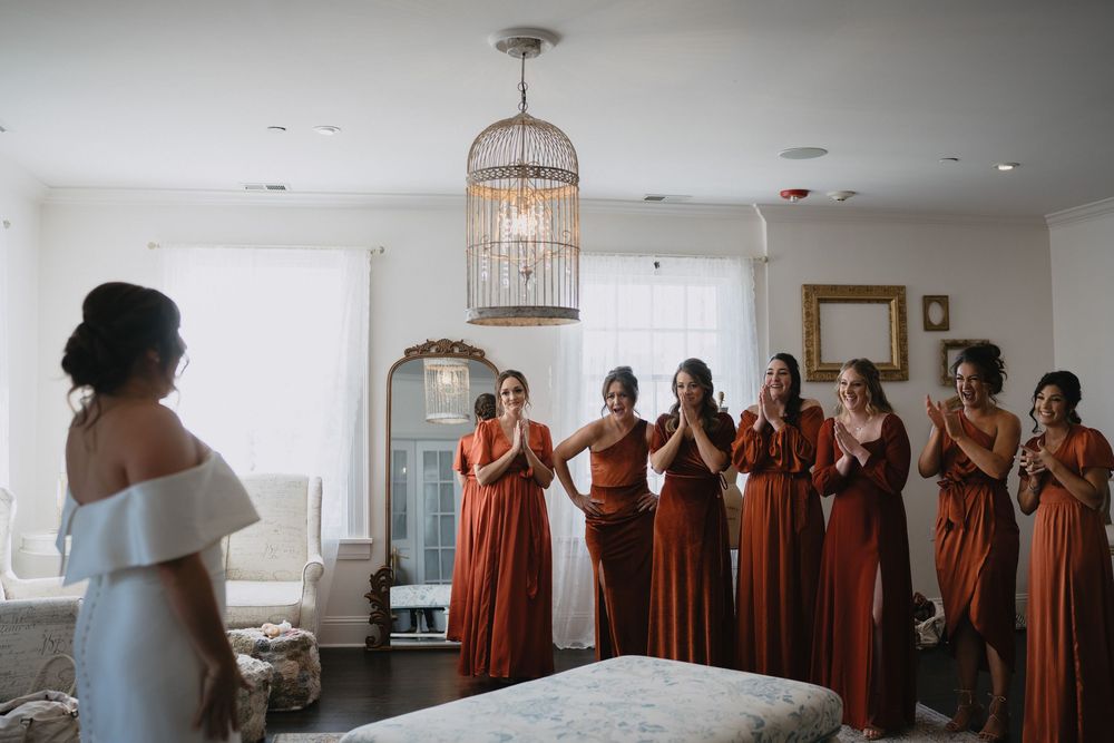 Bridesmaids in rust orange dresses react with joy as bride reveals her wedding gown in elegant bridal suite.