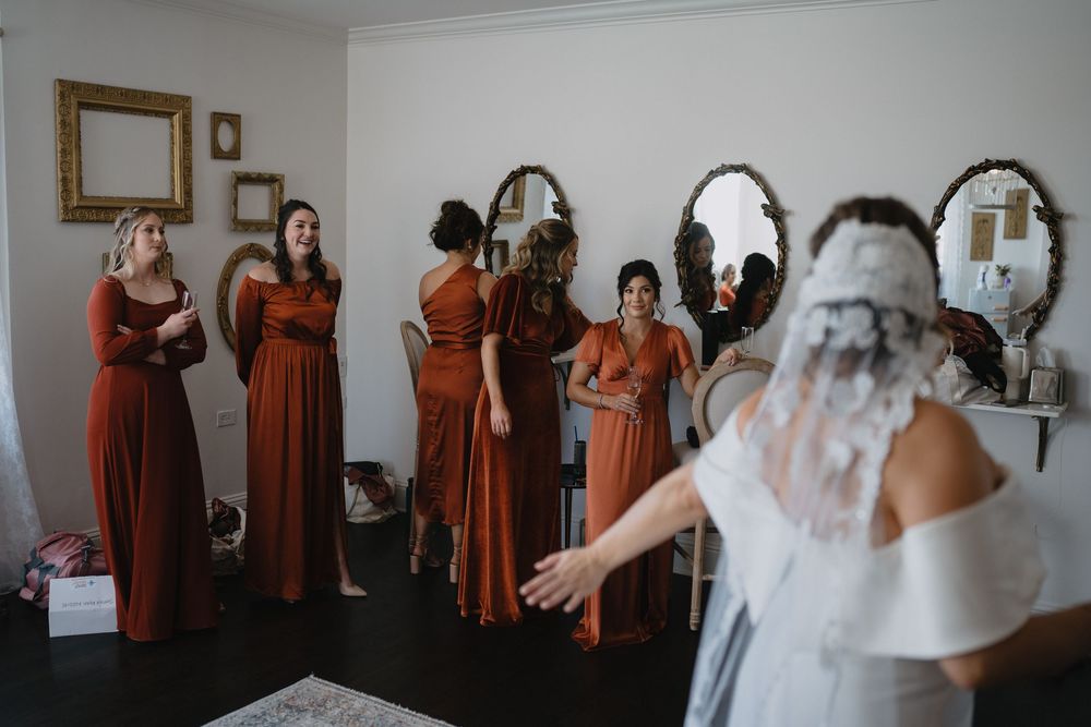 Bridesmaids in rust orange dresses react with joy as bride in white gown and veil turns to reveal her look.