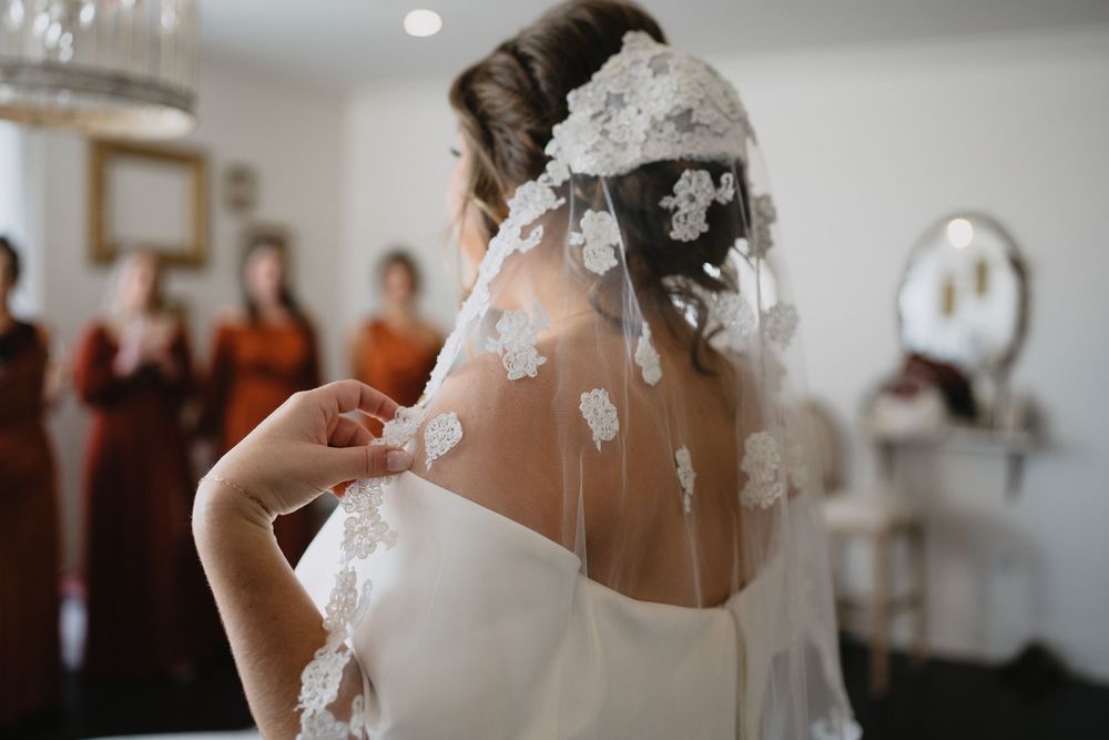 Bride in lace wedding dress and floral veil being buttoned up, bridesmaids in rust dresses visible in background.