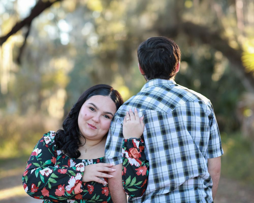 A couple in a plaid shirt and floral dress embrace during an outdoor photo session at sunset.