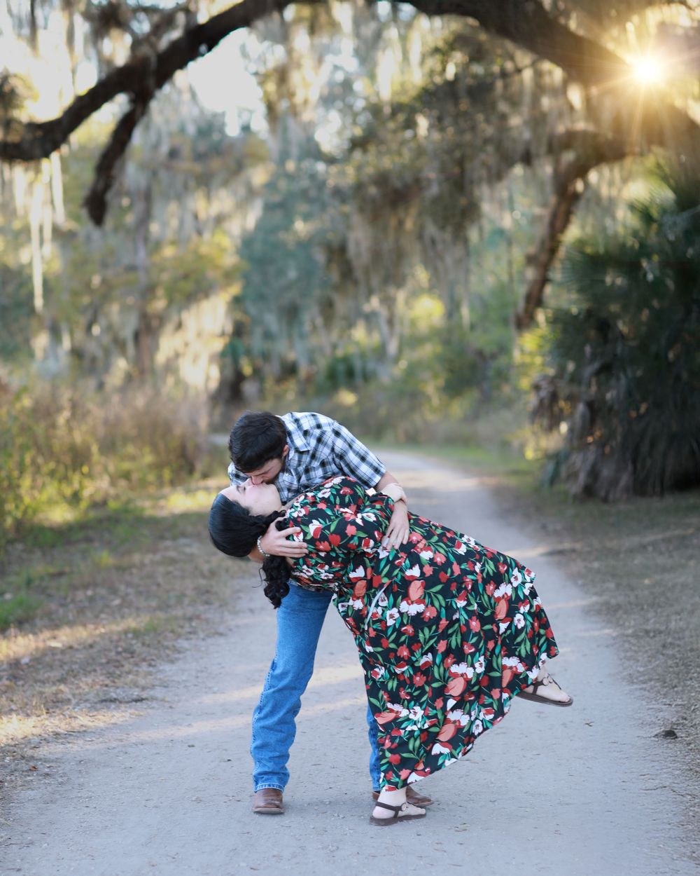 A person in a floral dress bends down to examine something along a tree-lined dirt path at sunset.