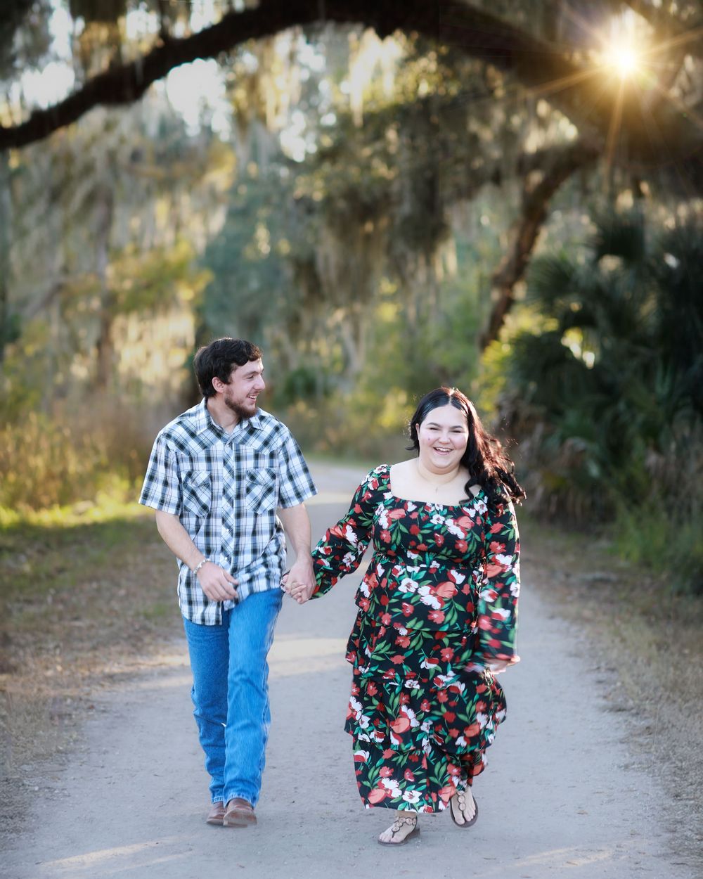 A couple in casual attire walks together on a sunlit dirt path through a wooded area.