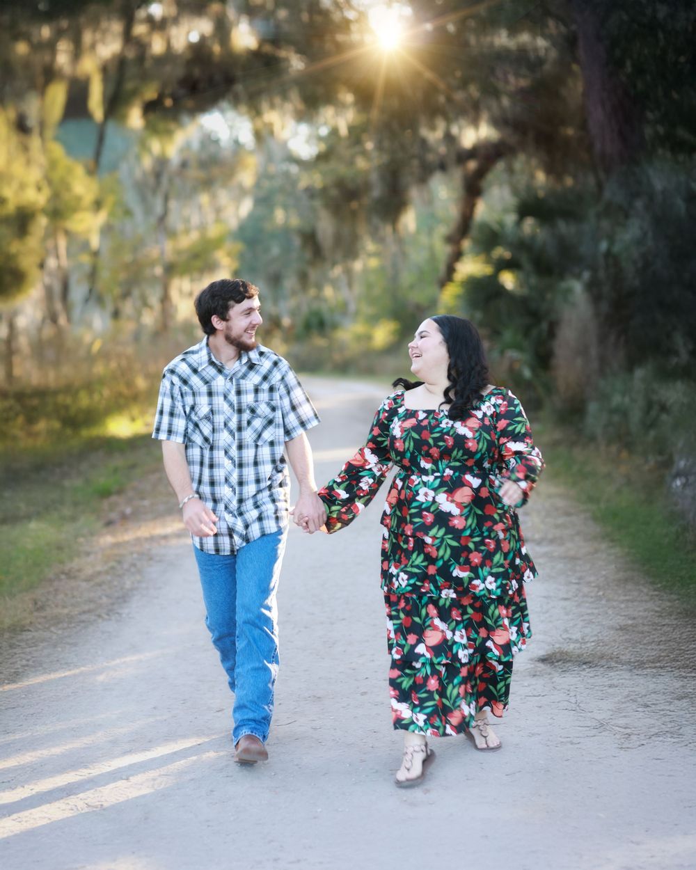 Two people walk together on a sunlit dirt path surrounded by trees with Spanish moss at sunset.