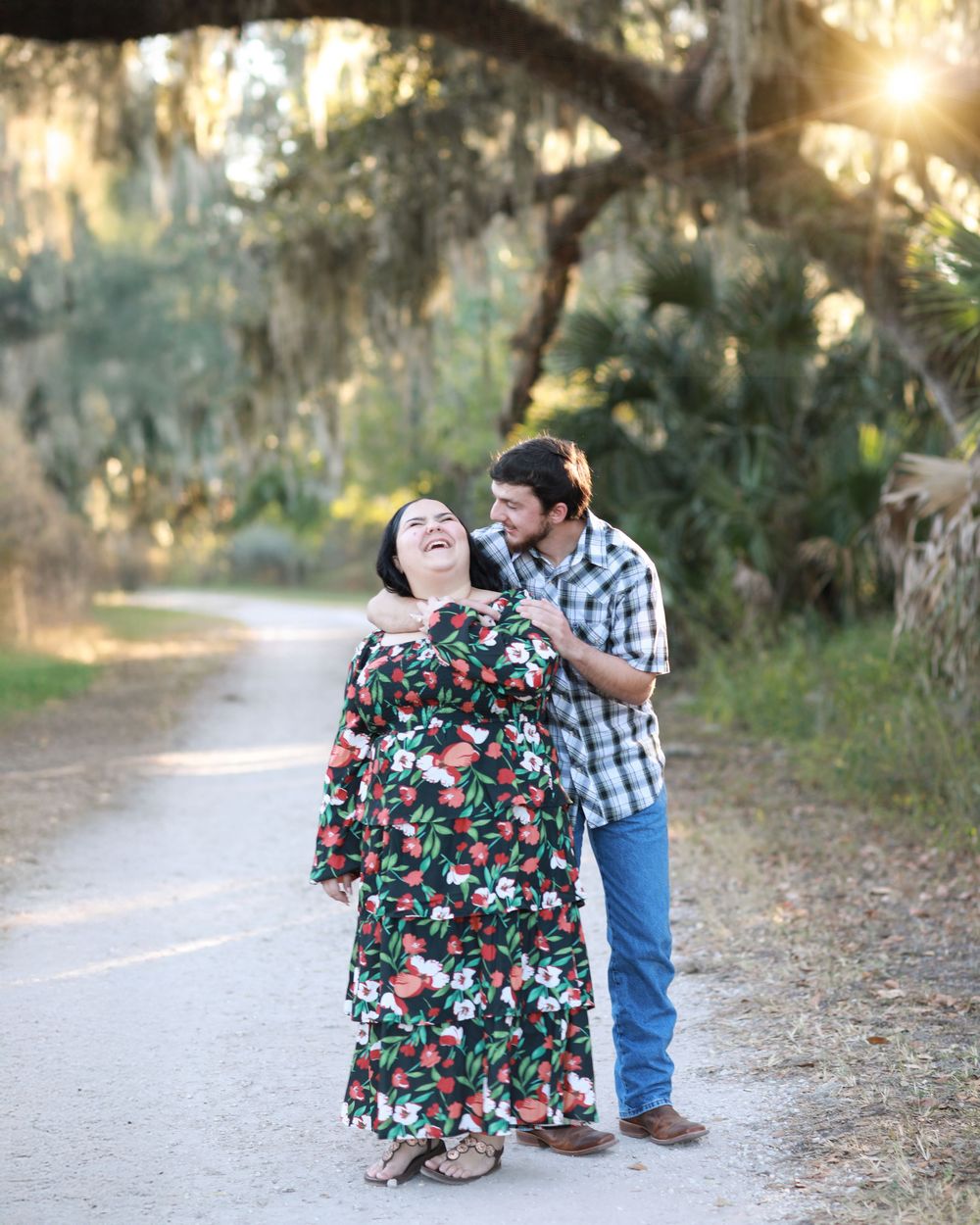 A couple laughs together on a nature trail with golden sunlight filtering through moss-draped trees.