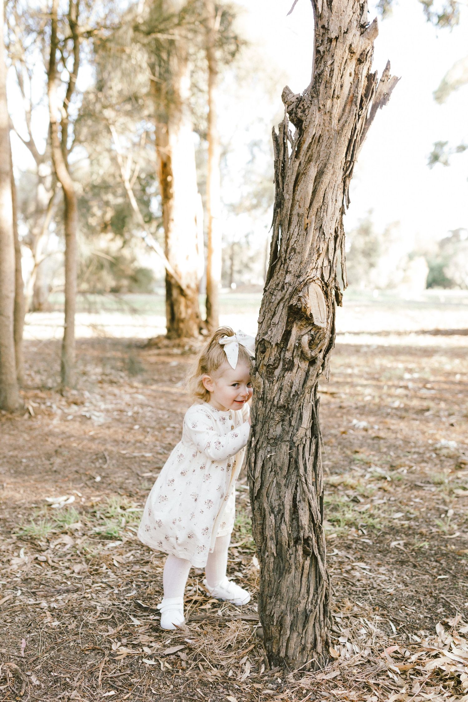 A small toddler in white leans against a tree trunk in a sunlit wooded area.