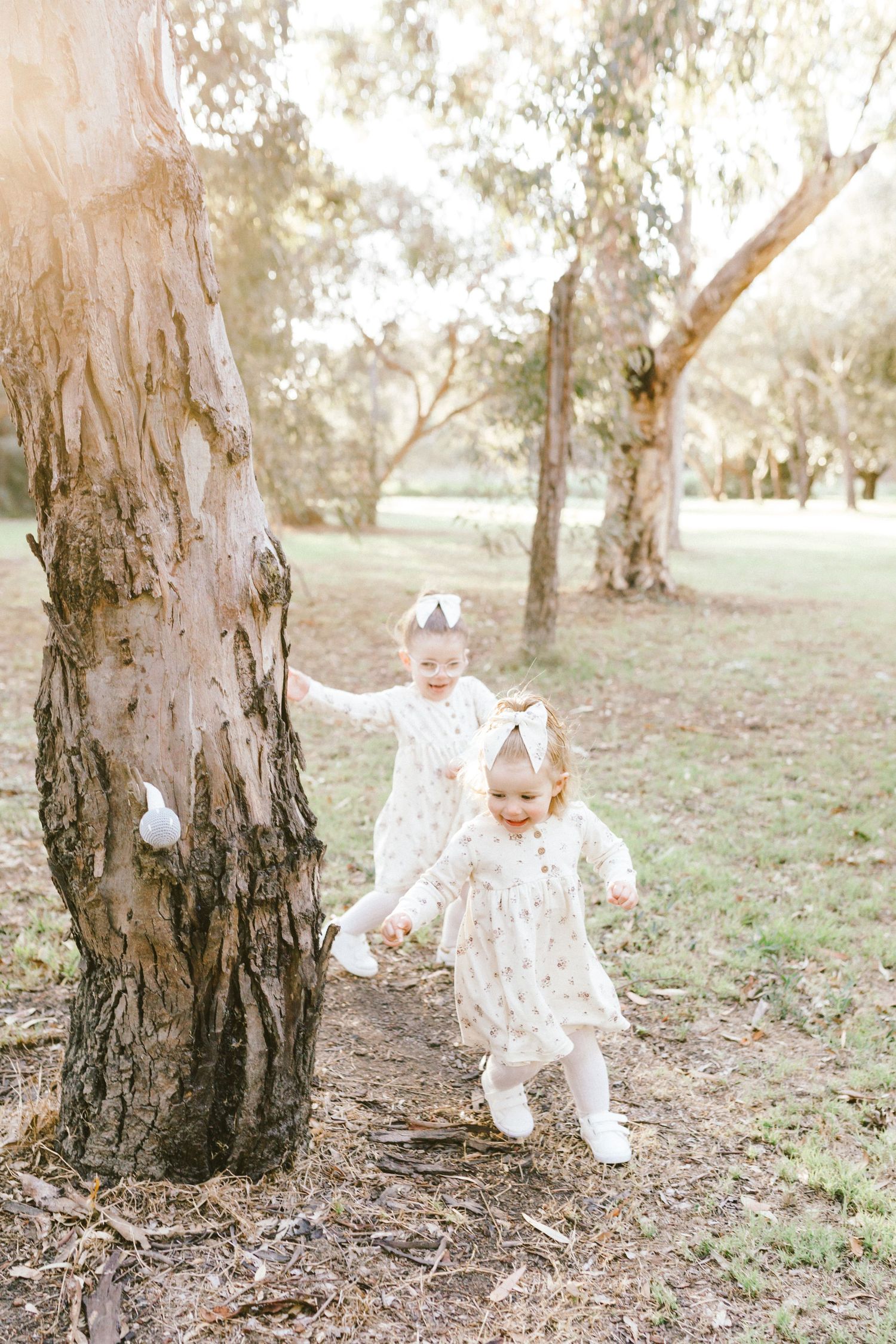 A toddler in a white lace outfit plays and explores around large tree trunks in a dreamy outdoor setting.