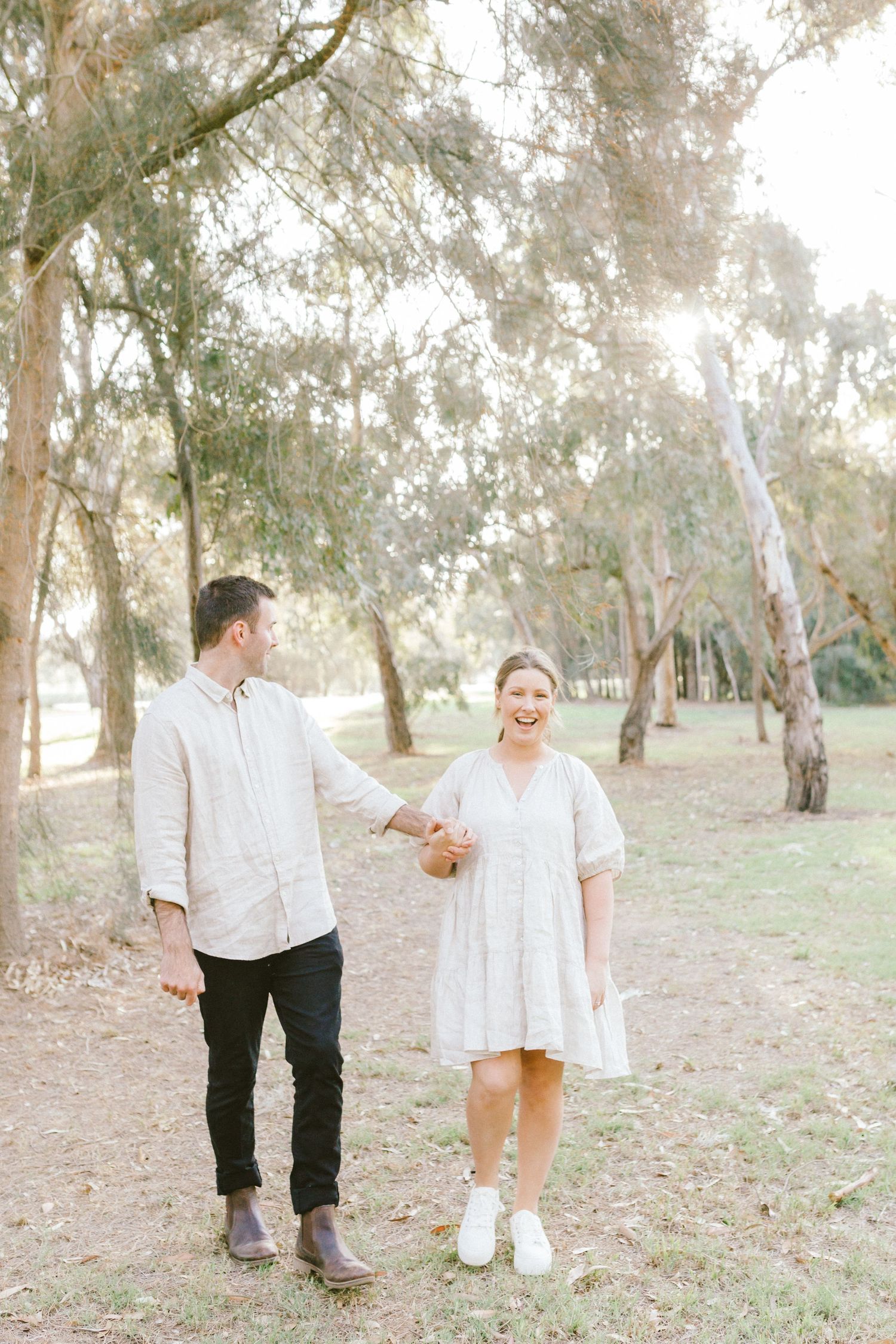 A couple in light colored clothing holding hands while standing together in a eucalyptus grove.