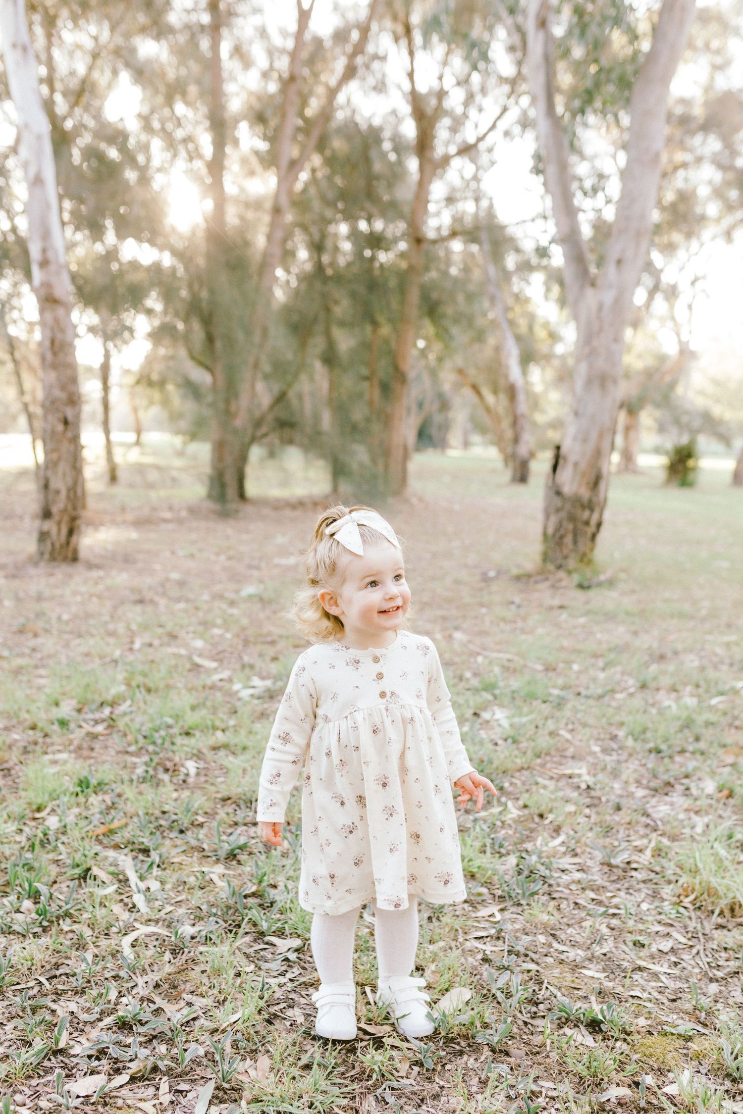 A young child in a floral dress stands among eucalyptus trees in a sunlit outdoor setting.