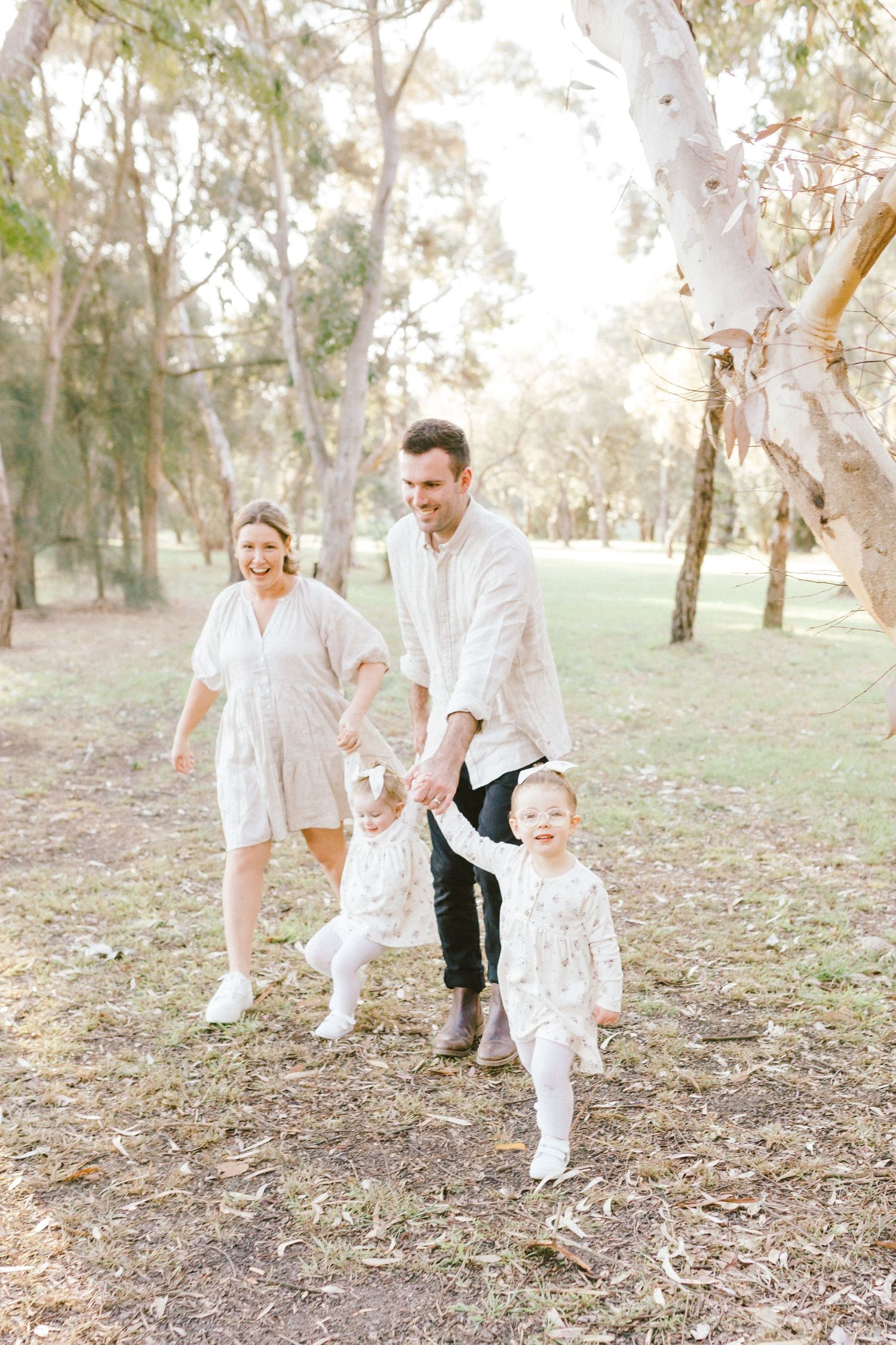 A family in white clothing walks and plays together on a wooded path during golden hour.