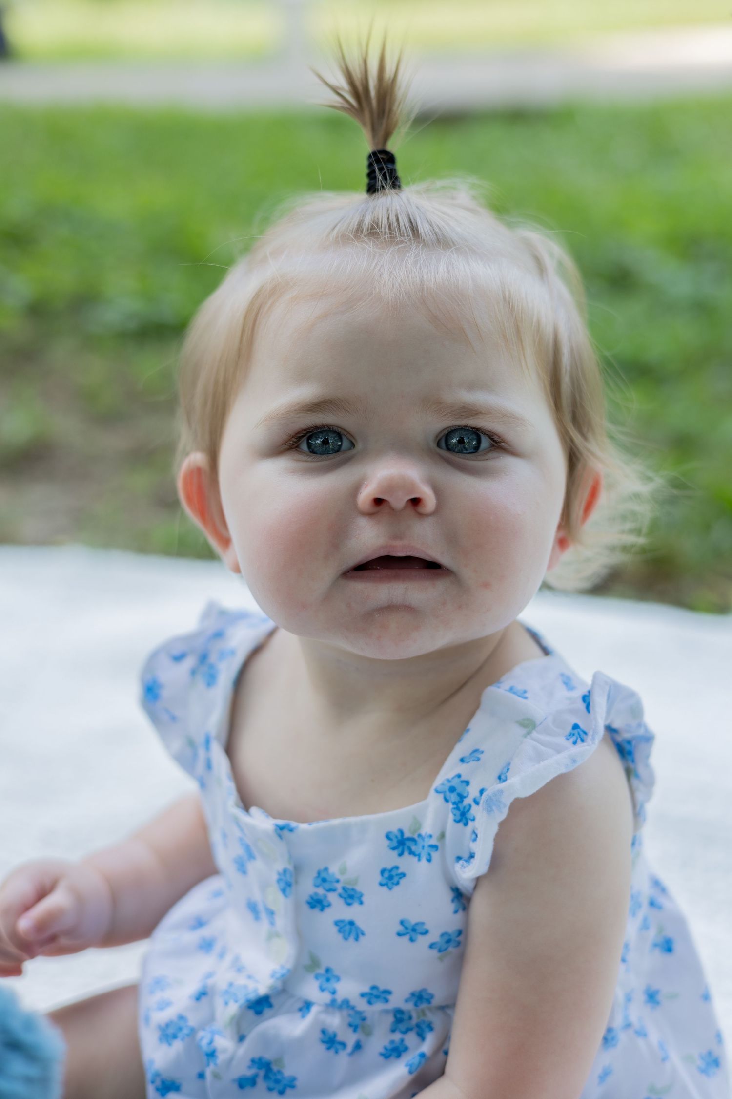 Baby in blue floral dress smiles sweetly at camera in outdoor setting.