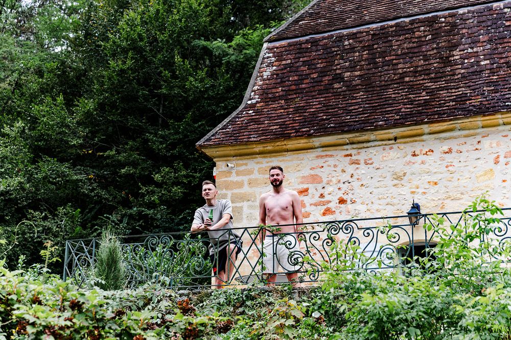 Two figures stand on a decorative balcony overlooking a lush garden at a rustic French country estate.