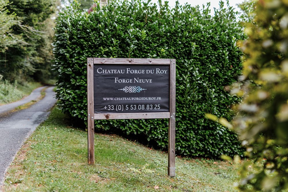 Wooden sign for Chateau Forge du Roy displayed on post near hedge-lined driveway.
