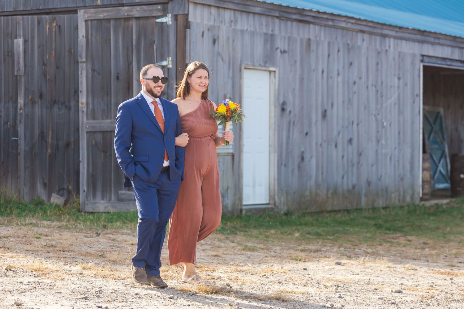 Couple in formal attire pose with sunflowers in front of rustic barn.