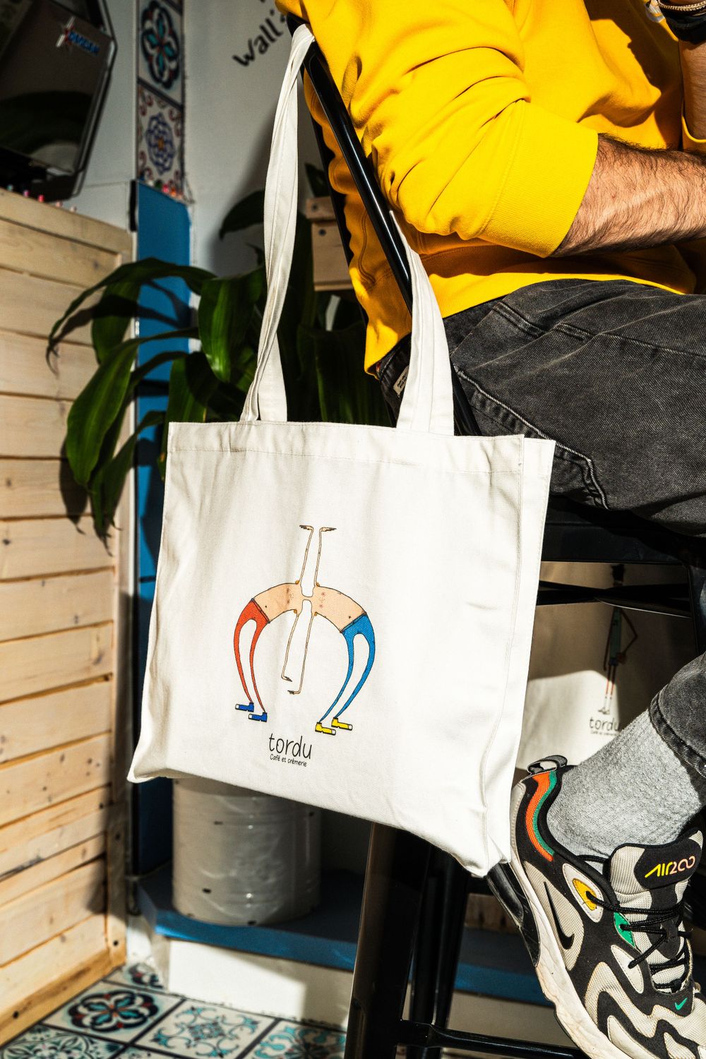 White tote bag with colorful geometric logo design displayed against wooden backdrop. Mtlshutter photography